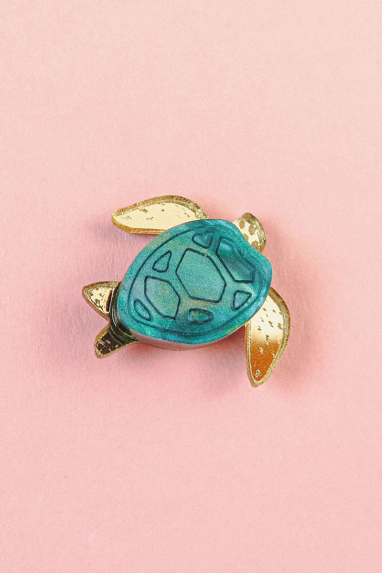 A small handcrafted sea turtle pin with a blue-green shell and gold-colored flippers, pictured on a pink background.