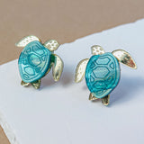 Sea Turtle Studs shaped like turtles with turquoise blue shells and gold-toned legs, heads, and fins bring a summery vibe, displayed on a white and beige background.