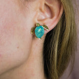 A close-up of a persons ear wearing gold and turquoise Sea Turtle Studs, with their light brown hair partially visible, captures an ocean-inspired, summery vibe.