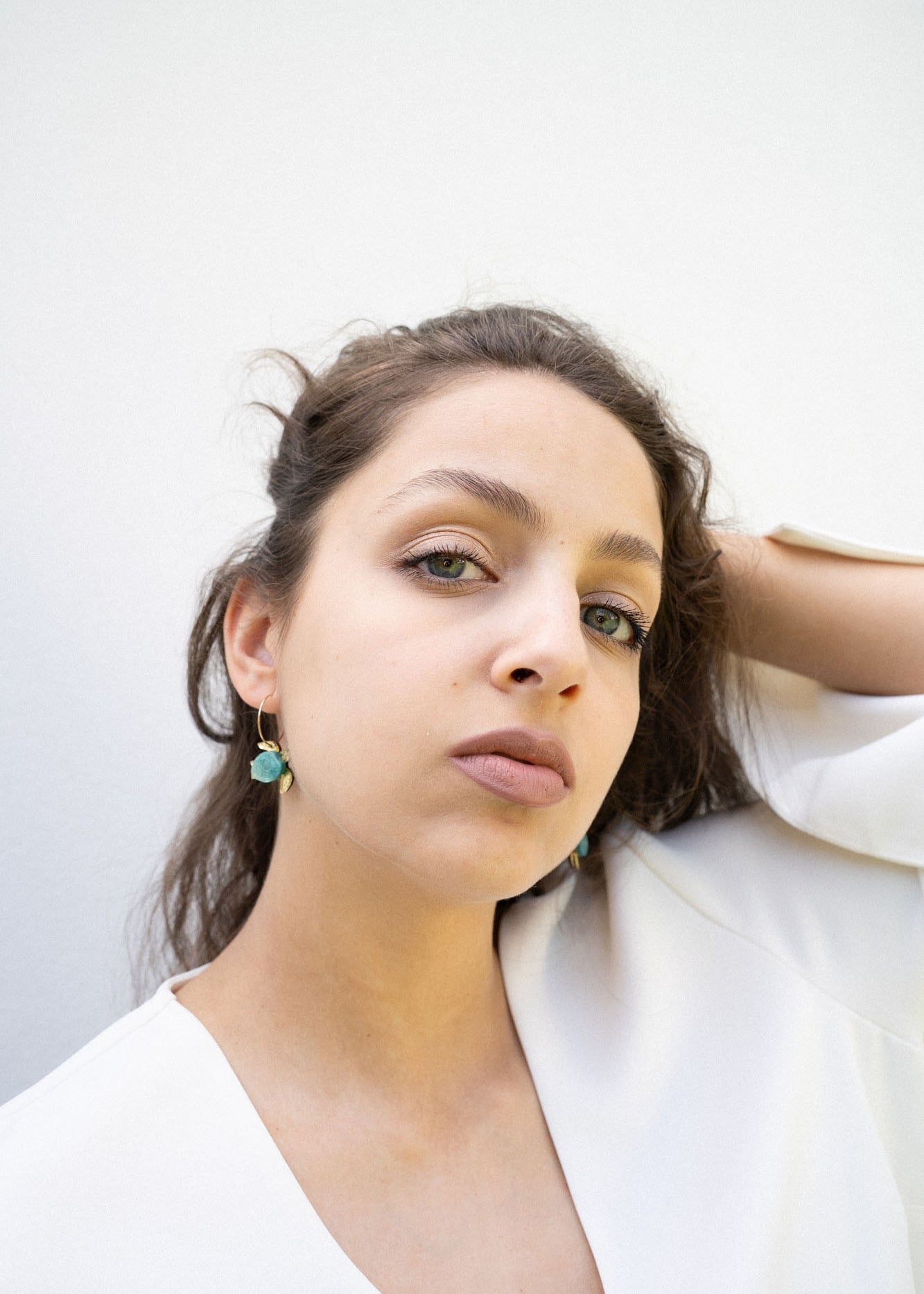 A young woman with light eyes and wavy brown hair poses against a plain white background, wearing a white top and handcrafted Little Turtle Hoop Earrings, with one hand touching her hair.