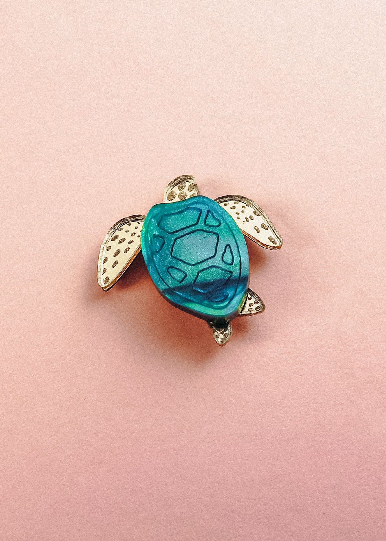 A small sea turtle pin, handcrafted with gold-colored flippers and a blue mirror-acrylic glass shell, rests on a light pink background.