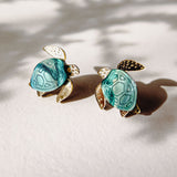 Two small Sea Turtle Studs with turquoise shells and gold-colored heads, legs, and tails rest on a light surface, casting soft shadows in natural light for a summery vibe.