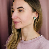A woman with straight, light brown hair and light skin wears a light pink top and turquoise Sea Turtle Studs, standing in front of a mauve curtain, gazing slightly to the left for a summery vibe.