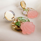 These handmade seashell earrings feature round gold studs, marbled green and cream discs, and pink translucent shell-shaped charms, all displayed on a soft beige fabric background. Lightweight and elegant for any occasion.