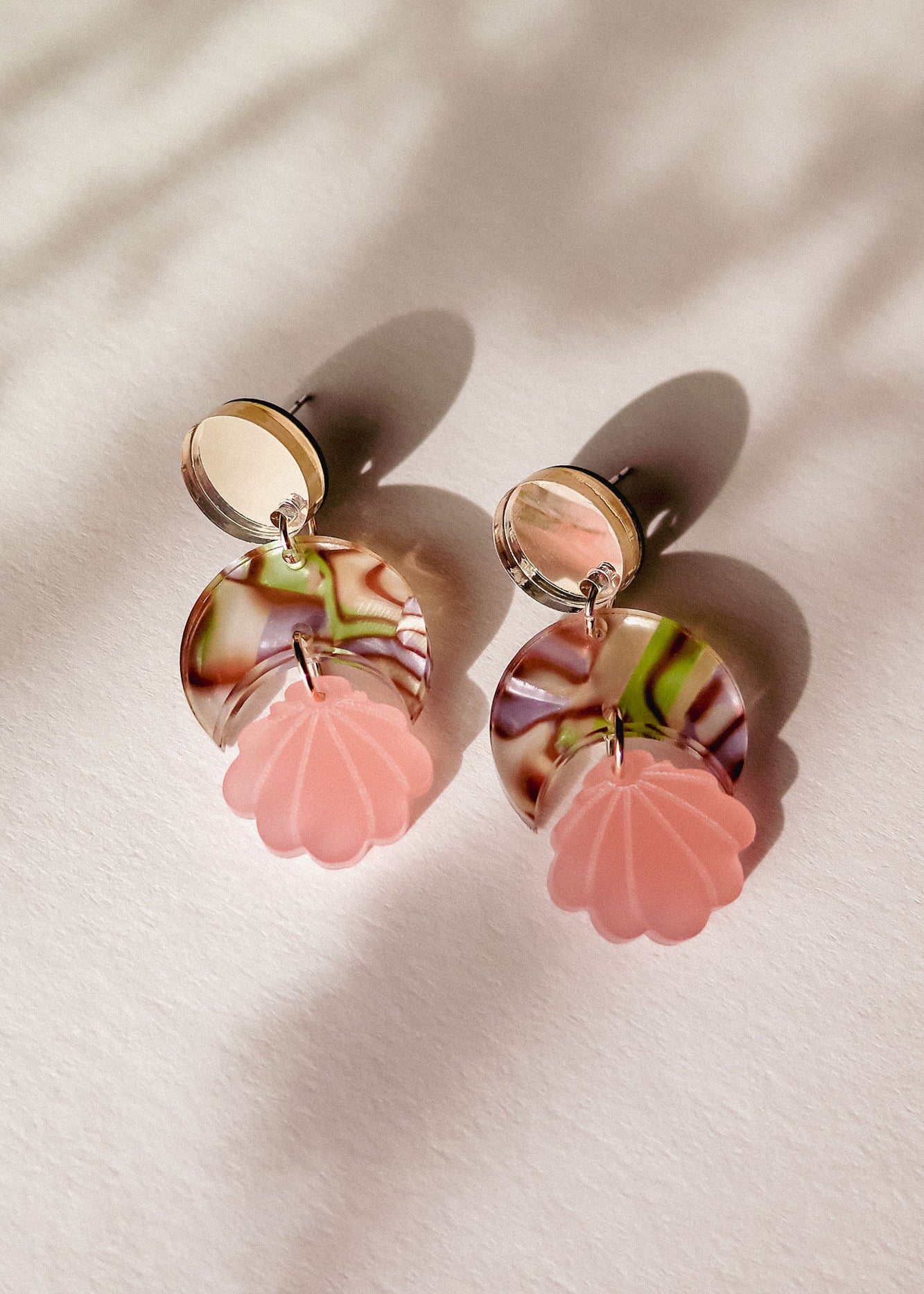 A pair of lightweight, handmade Seashell Earrings featuring round mirrored studs, marbled pink and brown circles, and pink shell-shaped pendants, displayed on a light surface with soft shadows.