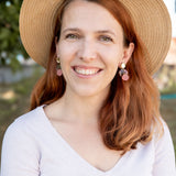 A woman with long auburn hair wearing a wide-brimmed straw hat, lightweight seashell earrings, and a light V-neck top smiles outdoors with greenery in the background.