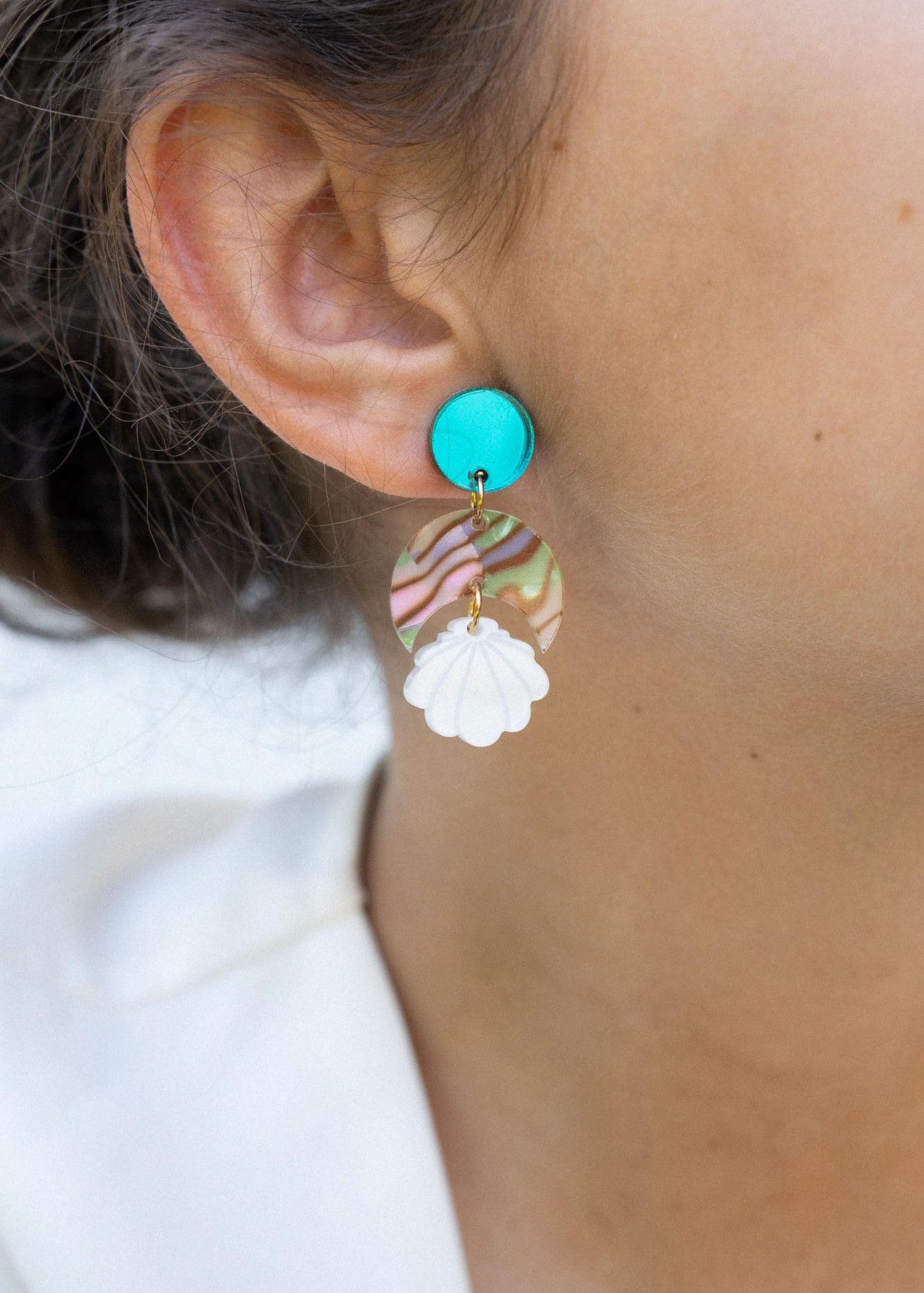 Close-up of a woman’s ear wearing a handmade, colorful earring with a turquoise stud, wavy striped section, and white floral-shaped dangle. Her hair is pulled back and she is wearing a white top. Lightweight for all-day comfort.