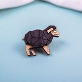 A wooden lapel pin shaped like a geometric ram with black and natural wood tones sits on a light blue surface, perfect for animal lovers and fans of polygon design, with white fabric in the background.