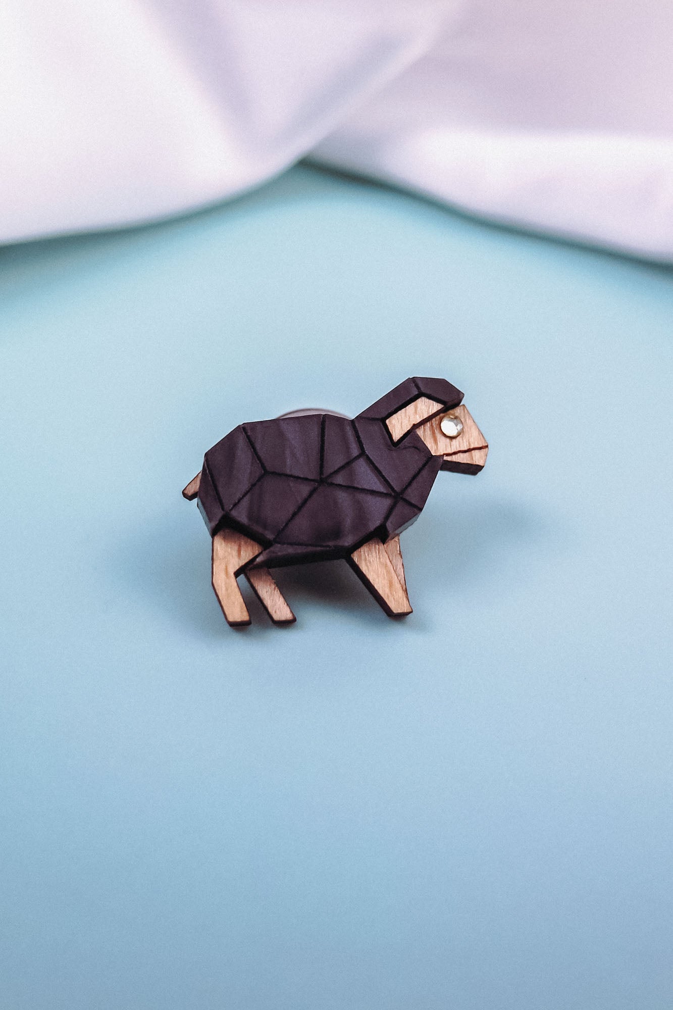 A wooden lapel pin shaped like a geometric ram with black and natural wood tones sits on a light blue surface, perfect for animal lovers and fans of polygon design, with white fabric in the background.