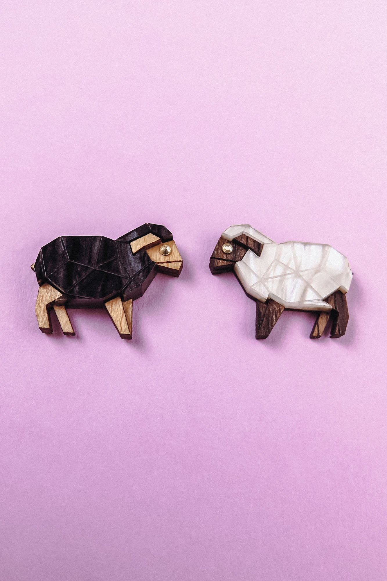 Two small wooden Sheep Badges shaped like sheep, one dark brown and one white, placed on a light purple background—a charming accessory choice for animal lovers.