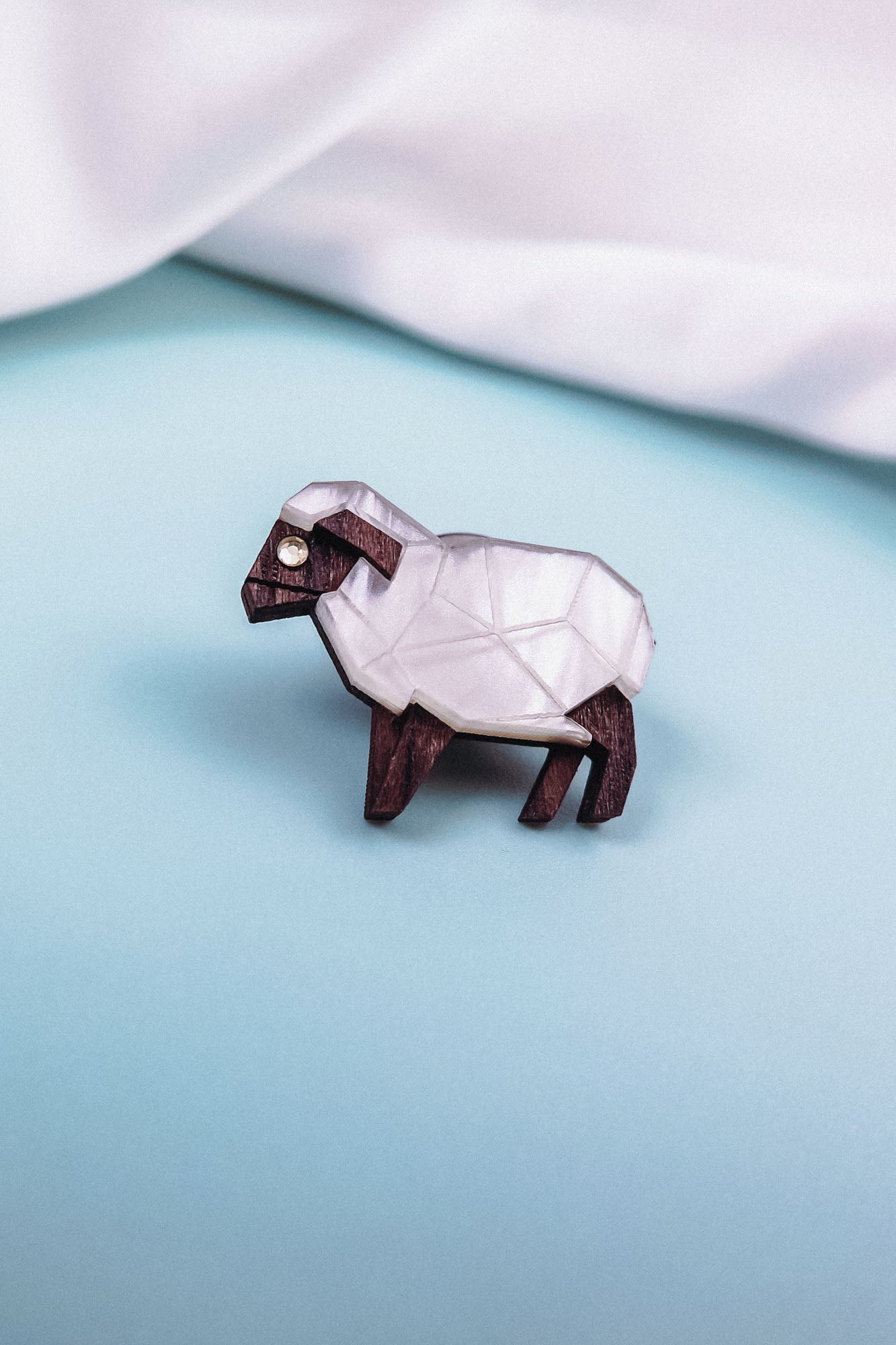A small, geometric sheep figurine with a white, faceted body and dark brown legs and head, placed on a light blue surface—perfect inspiration for an animal lovers’ Sheep Badge or decorative lapel pin.