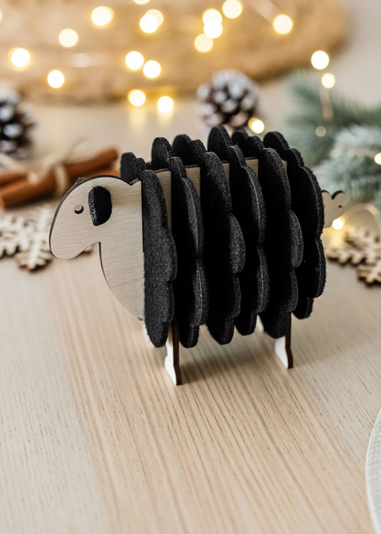 A small wooden sheep figurine made with black felt adds charm to your home decor, standing on a light wooden table with festive lights, pinecones, and greenery blurred in the background.