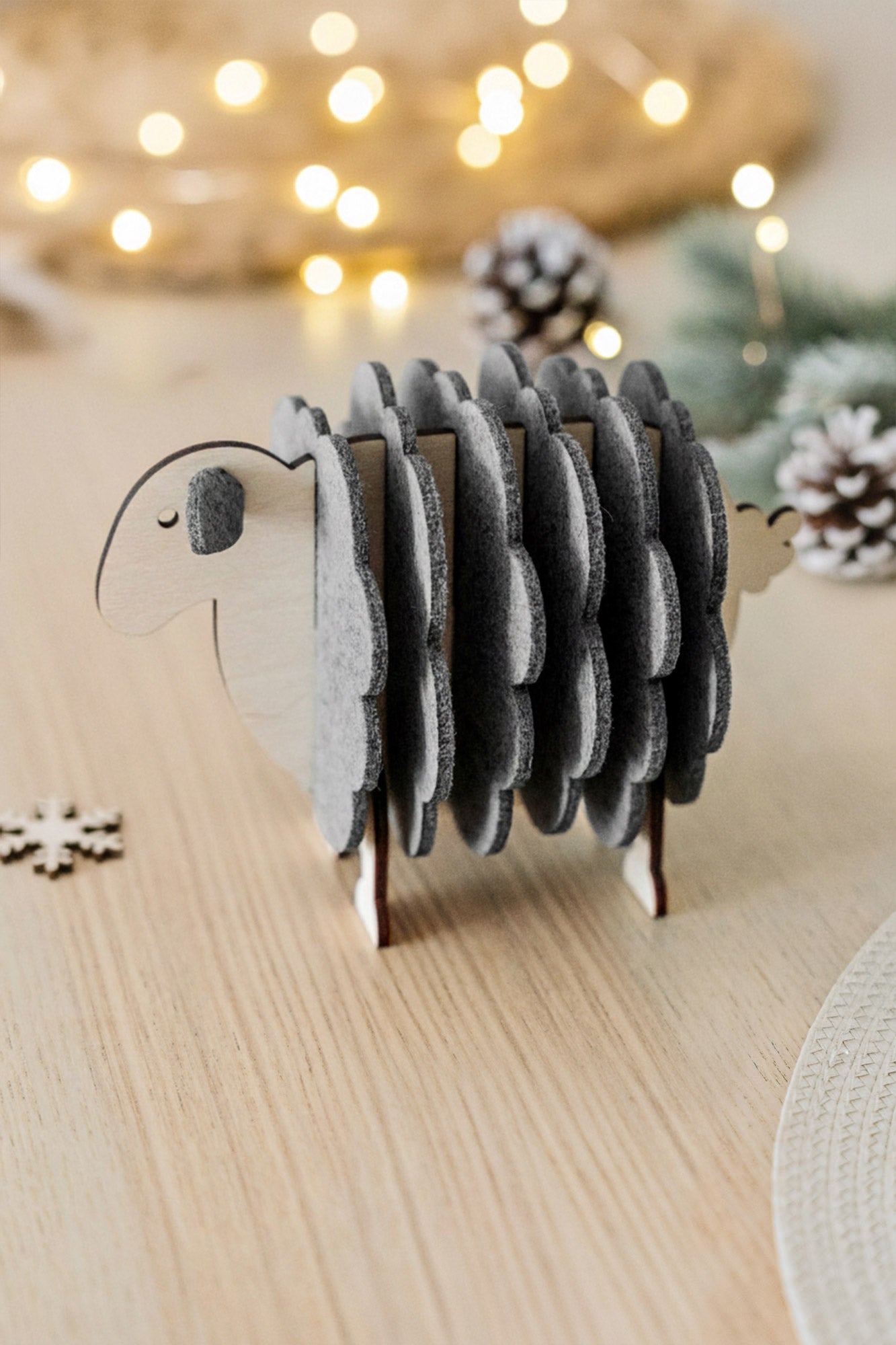 A wooden sheep-shaped coaster holder with felt coasters sits on a light wooden table, surrounded by pinecones, a snowflake decoration, and softly glowing fairy lights—charming home decor for cozy gatherings.