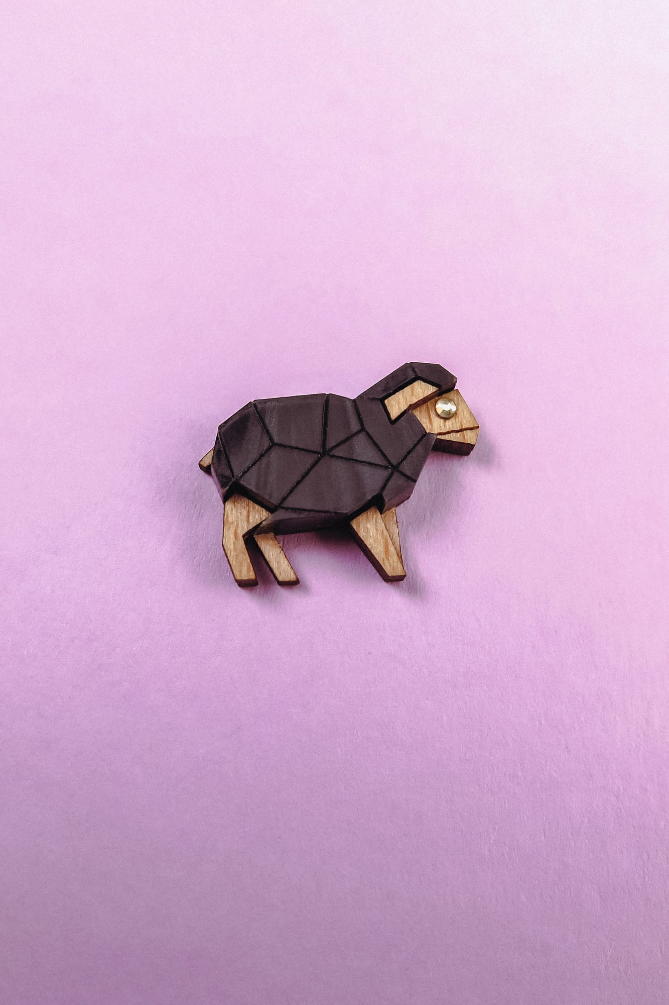 A small, geometric goat figurine with a polygon design, crafted from wood and dark material, sits on a pastel pink background. The minimalist piece features angular shapes—perfect for animal lovers with a flair for modern decor.
