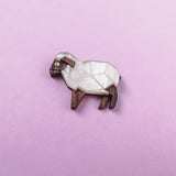 A small, geometric sheep figurine made of wood and white material sits on a pastel pink background—an ideal Sheep Badge or keepsake for animal lovers.