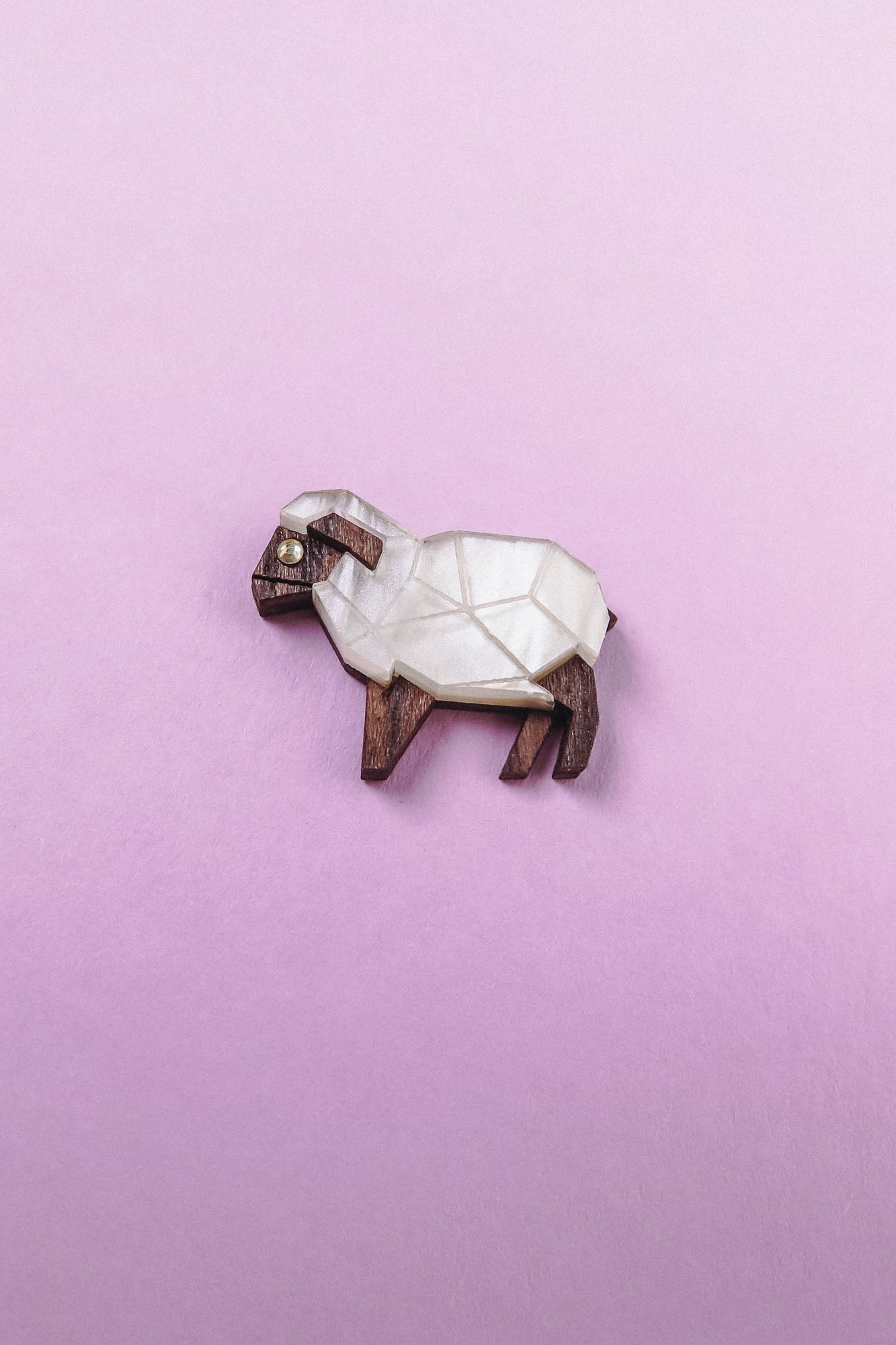 A small, geometric sheep figurine made of wood and white material sits on a pastel pink background—an ideal Sheep Badge or keepsake for animal lovers.