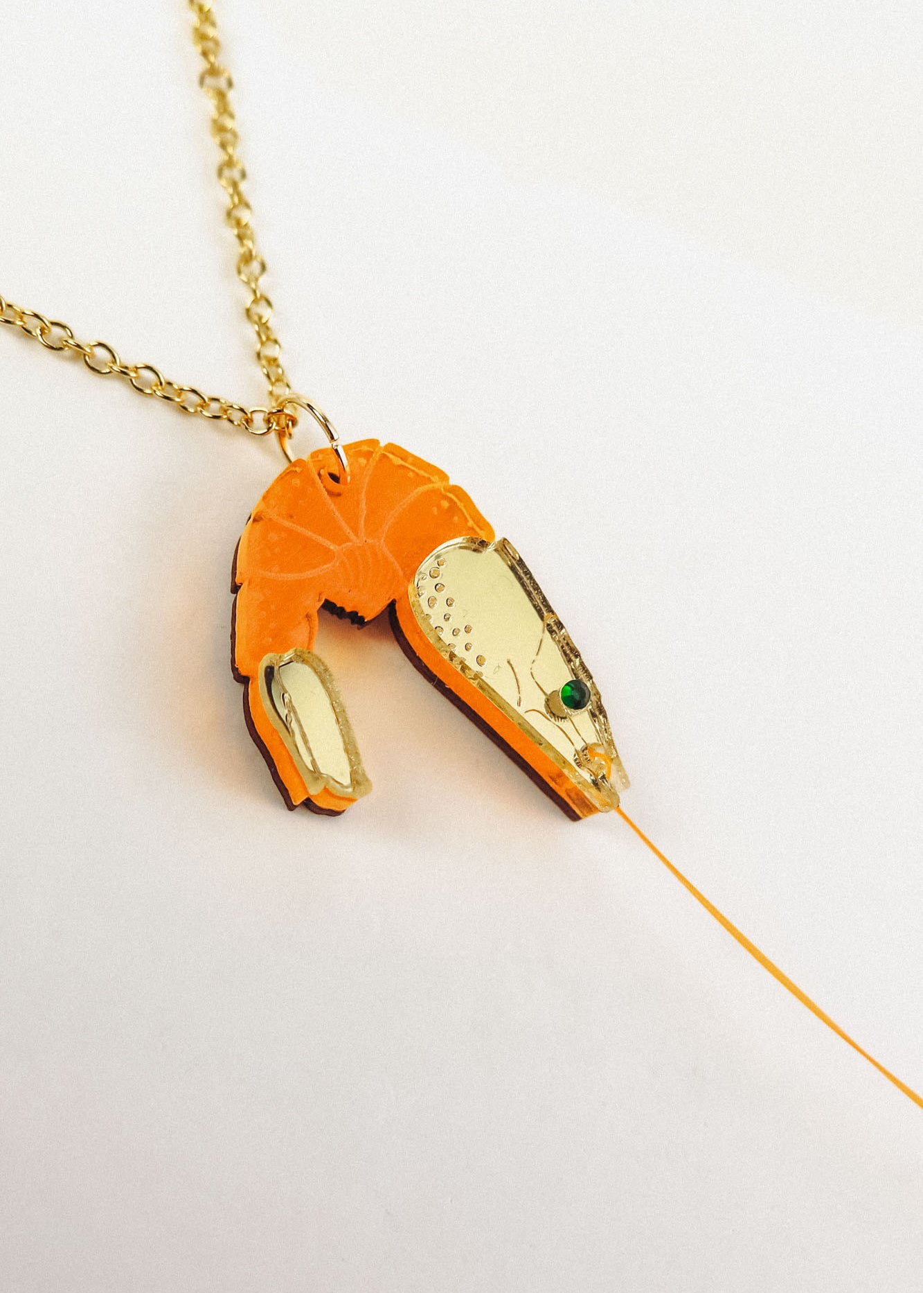 A playful Shrimp Pendant Necklace with a gold chain features an orange shrimp pendant adorned with gold accents and a green gem eye, creating a whimsical touch against a white background.