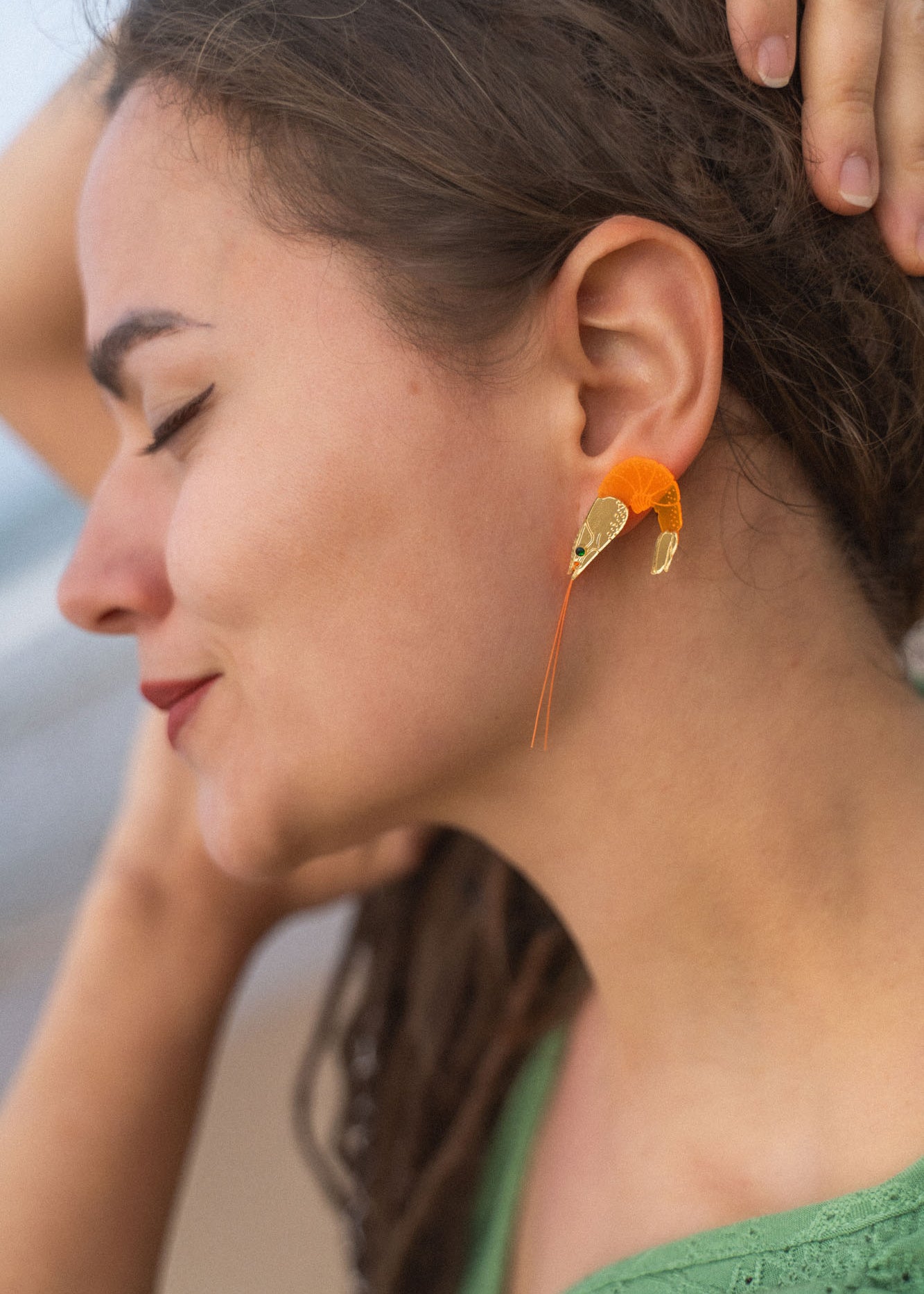 A woman with closed eyes and a relaxed expression wears Shrimp Stud earrings accented with emerald rhinestones. Outdoors, she touches her hair and is dressed in a green top.