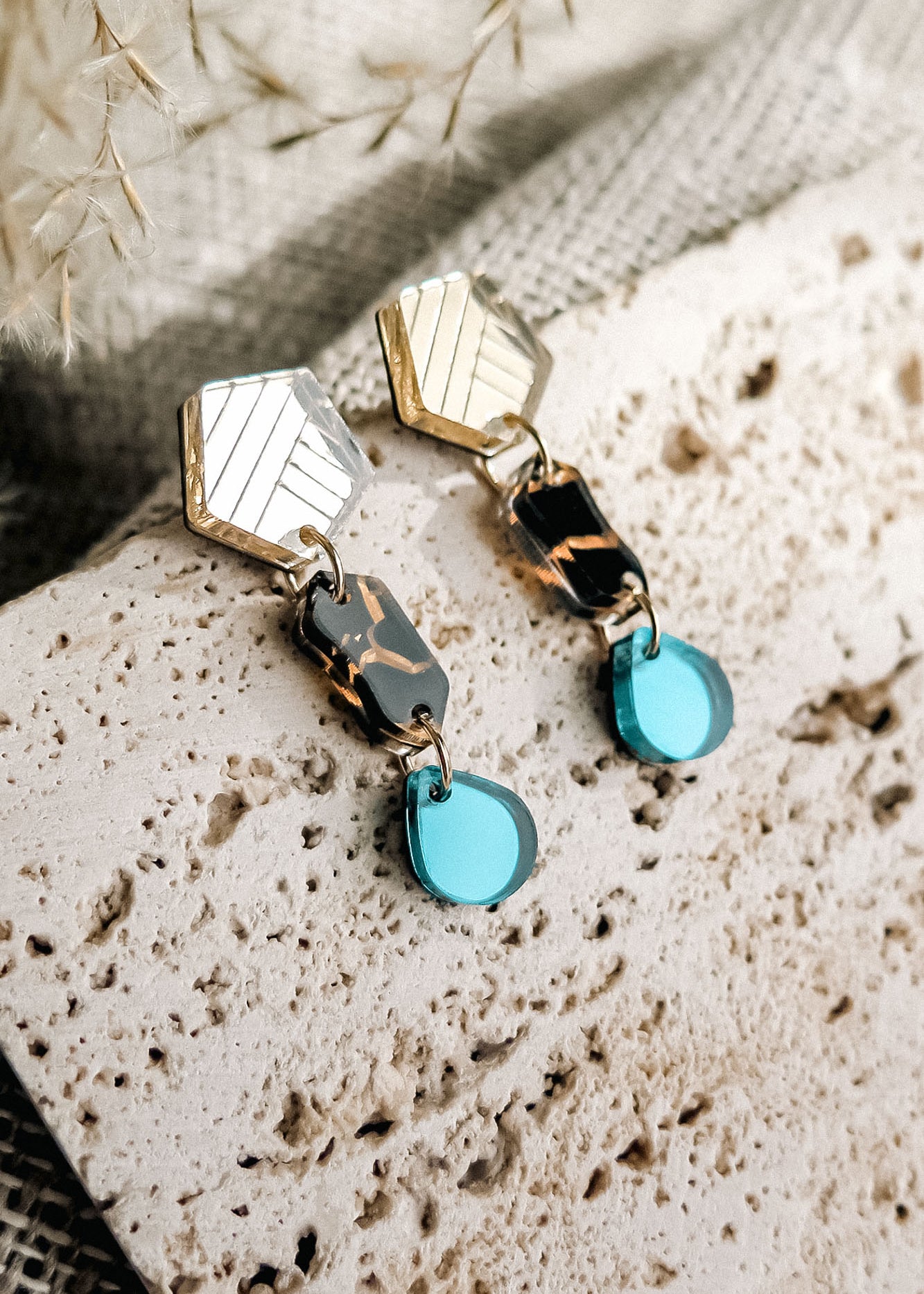 A pair of handcrafted geometric drop earrings with gold, tortoiseshell, and turquoise blue elements rest on a textured stone surface with a neutral fabric and dried plants in the background.