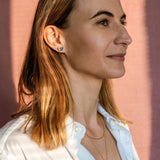 A woman with straight, shoulder-length light brown hair, wearing a white shirt, layered gold necklaces, and tiny heart studs, stands in front of a pink background, looking to the right with a calm expression.