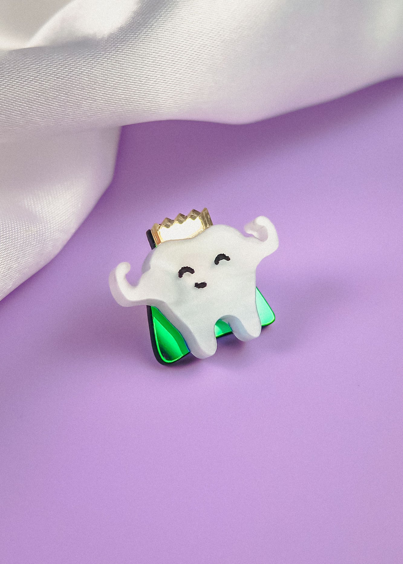 A cute Tooth King Pin shaped like a smiling tooth with a gold crown and flexed arms sits on a shiny green base, placed on a purple surface with white fabric in the background—a charming handcrafted accessory for any collection.
