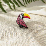 A handcrafted, colorful toucan pin with a yellow beak and red and black body rests on textured beige fabric, surrounded by green fern leaves and soft white cloth in the background.
