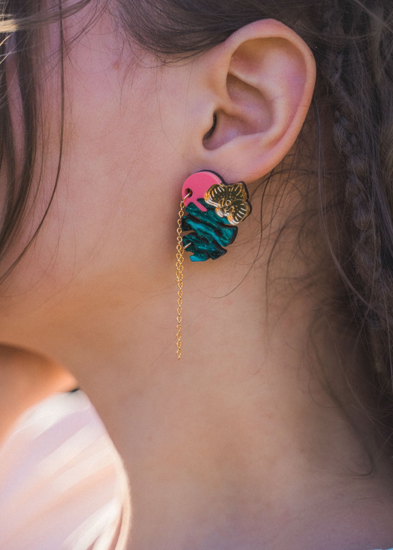 Close-up of a person’s ear wearing Tropicana Earrings—a tropical-inspired design with a gold tiger head, blue and pink details, and a delicate gold chain. The person’s hair is partially braided.