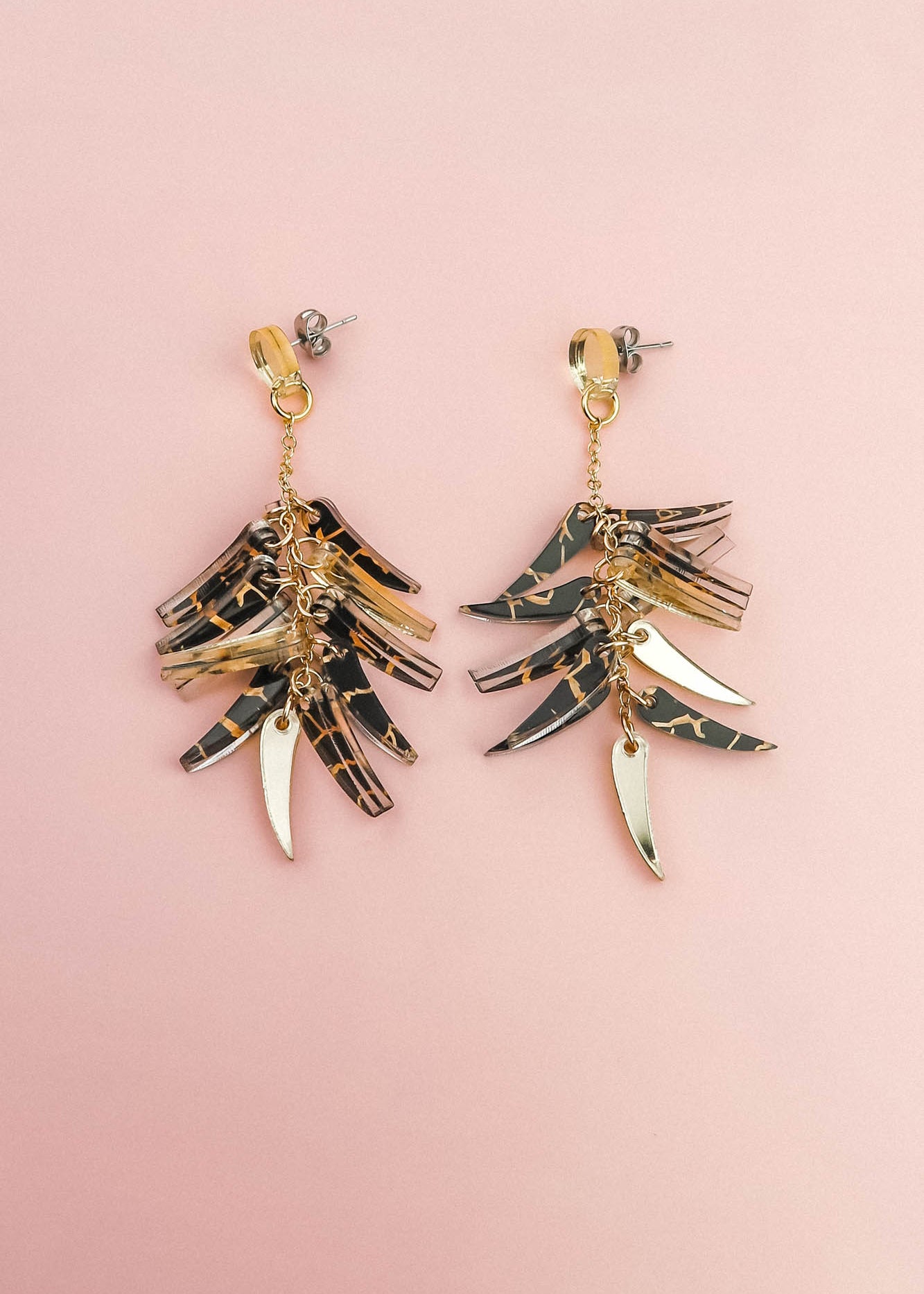 A pair of featherlight dangle earrings with layered gold and tortoiseshell-patterned pieces arranged in a fan-like design, displayed on a light pink background.