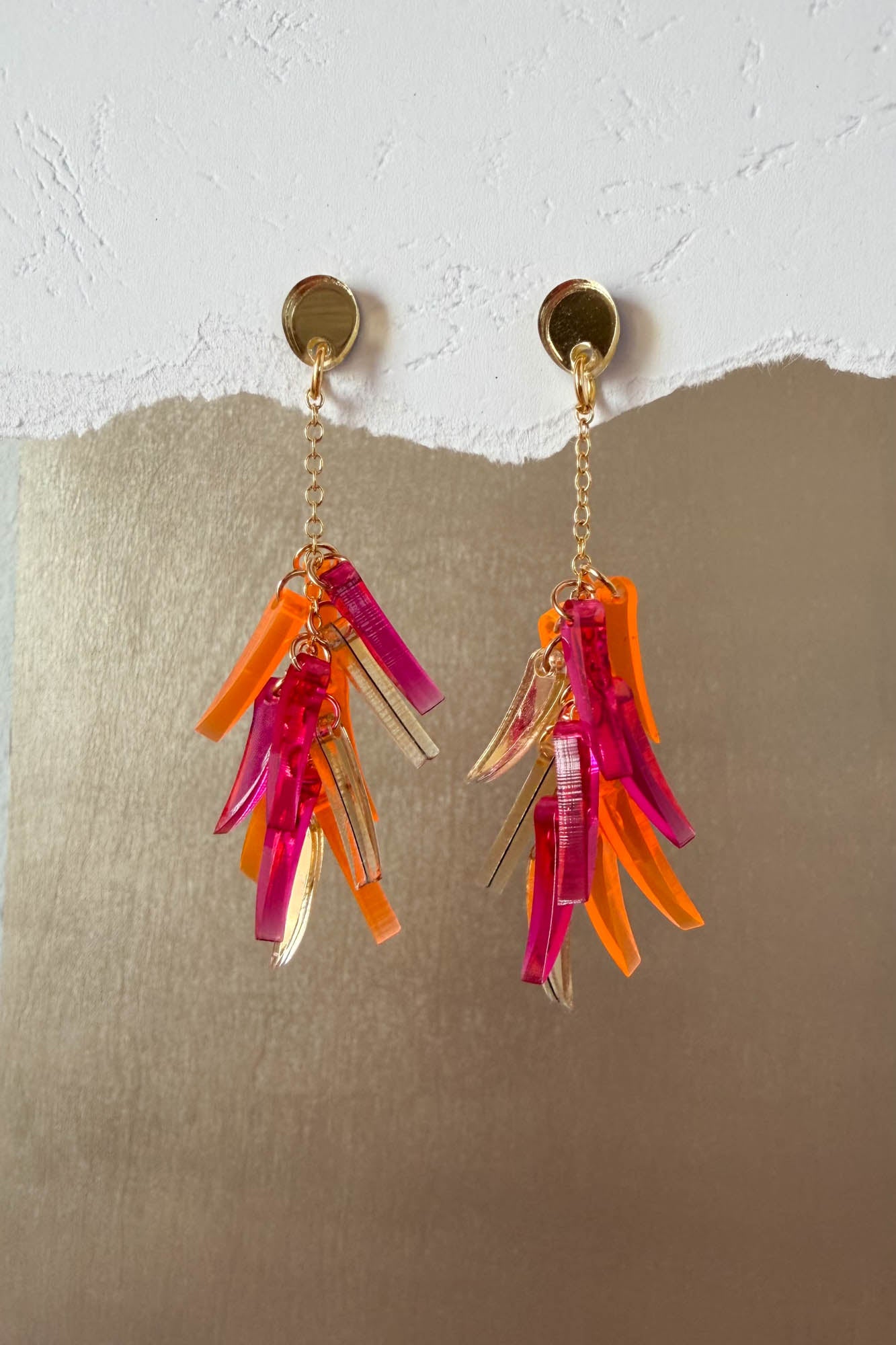 A pair of featherlight dangle earrings with gold studs and chains, each featuring clusters of translucent pink, orange, and clear acrylic shards, displayed against a textured white and metallic gold background.