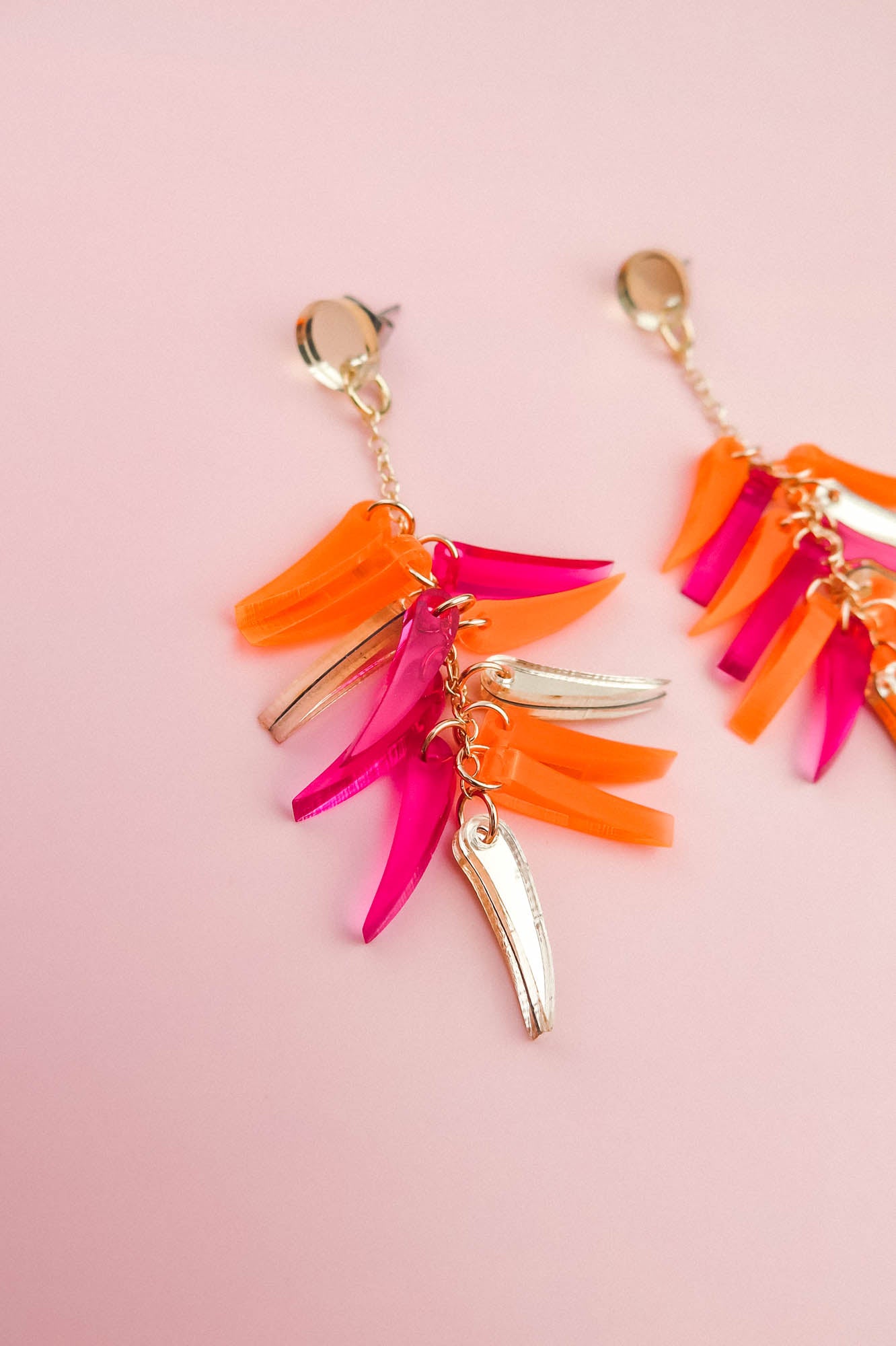 A pair of featherlight dangle earrings on a pink background, featuring gold metal accents and colorful acrylic charms in orange, pink, and gold.