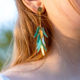 Close-up of a person wearing featherlight gold stud dangle earrings with metallic and turquoise leaf-shaped charms. The person has light brown hair, fair skin, and is dressed in a white top.