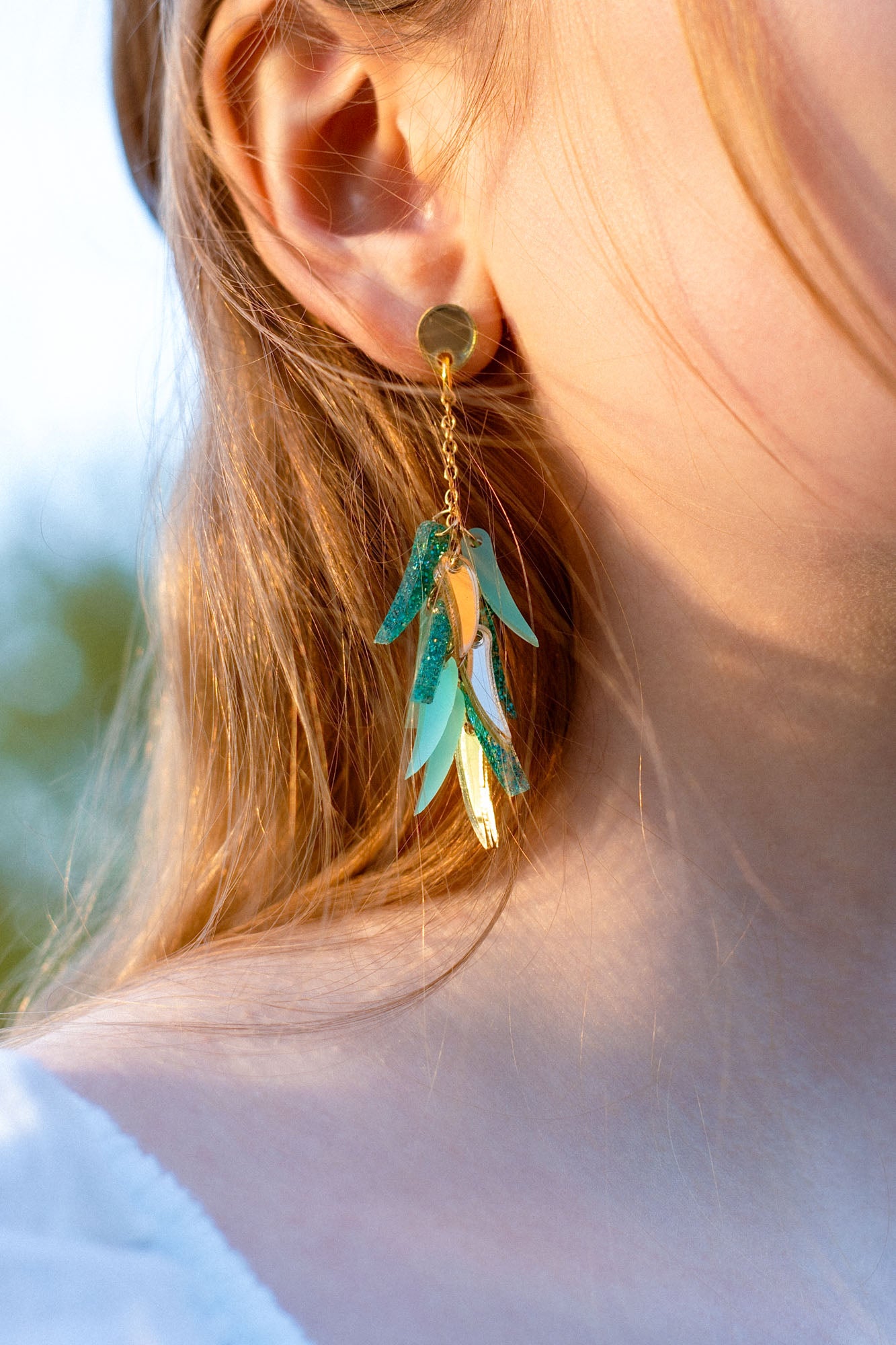 Close-up of a person wearing featherlight gold stud dangle earrings with metallic and turquoise leaf-shaped charms. The person has light brown hair, fair skin, and is dressed in a white top.
