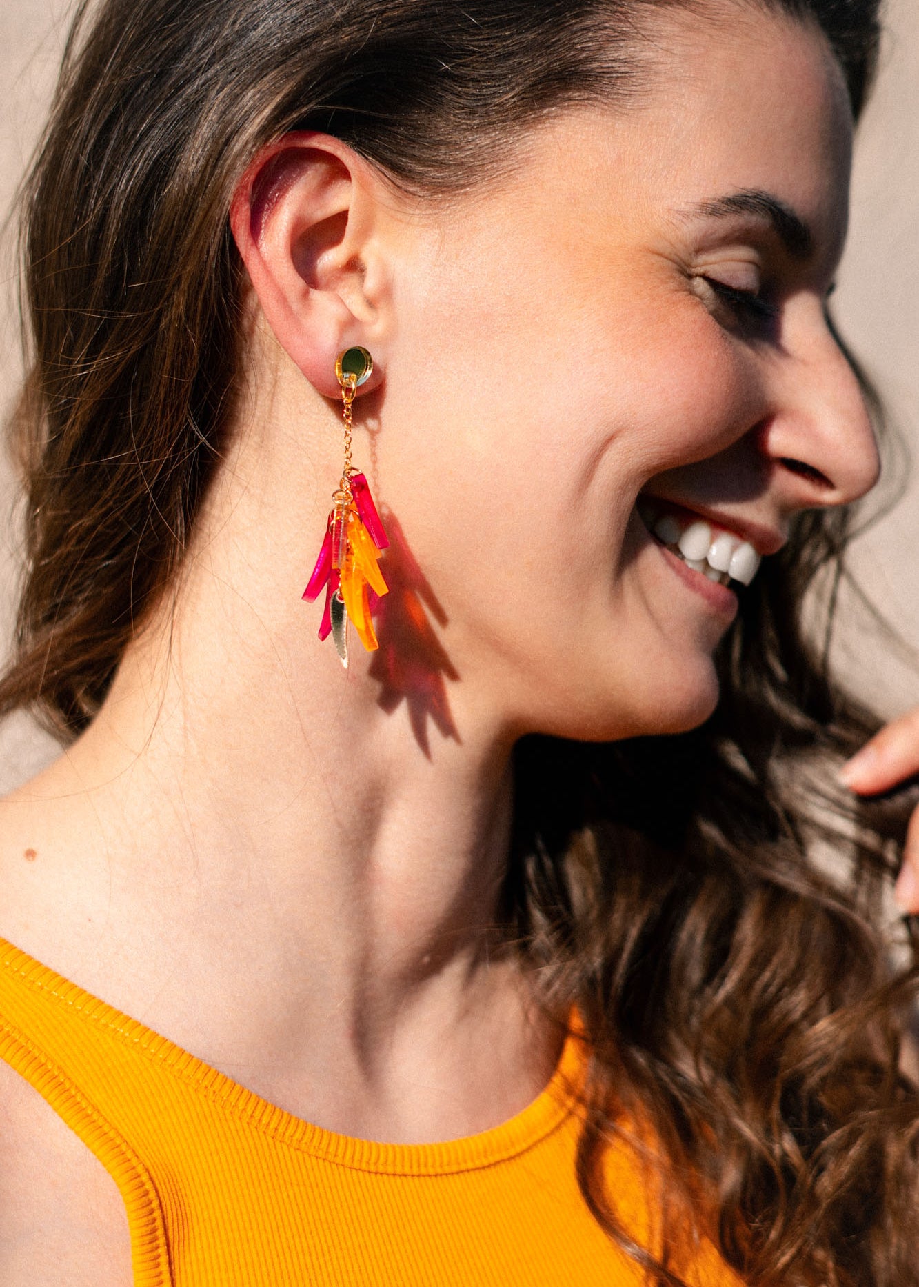 A woman with long brown hair smiles while wearing a sleeveless orange top and featherlight dangle earrings with pink, red, and yellow accents. The image highlights her earrings and natural makeup.