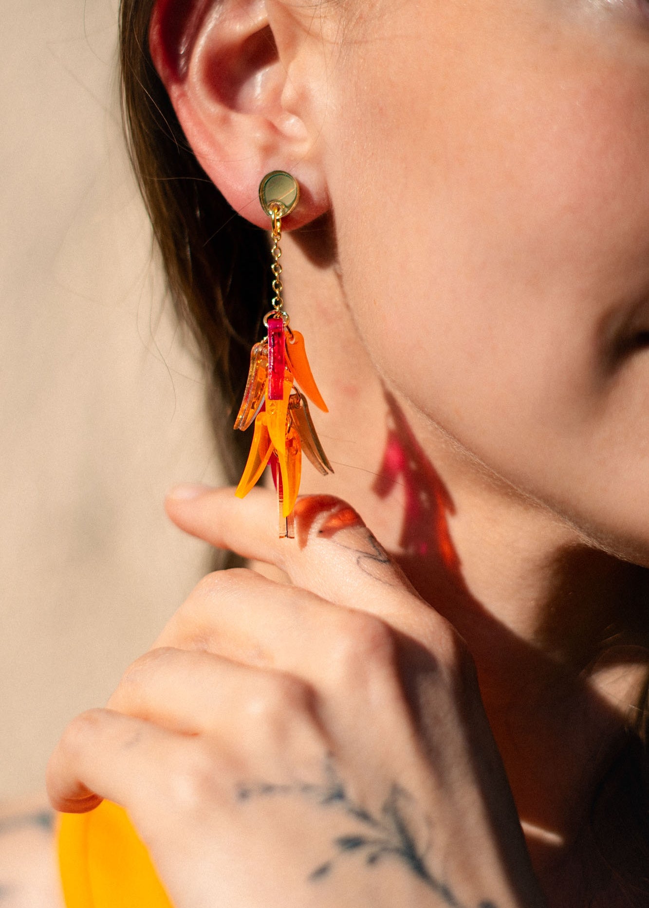 Close-up of a woman wearing featherlight dangle earrings shaped like orange flower petals. She is smiling, and her hand with a delicate tattoo touches her neck near the earring. Her hair is tucked behind her ear.
