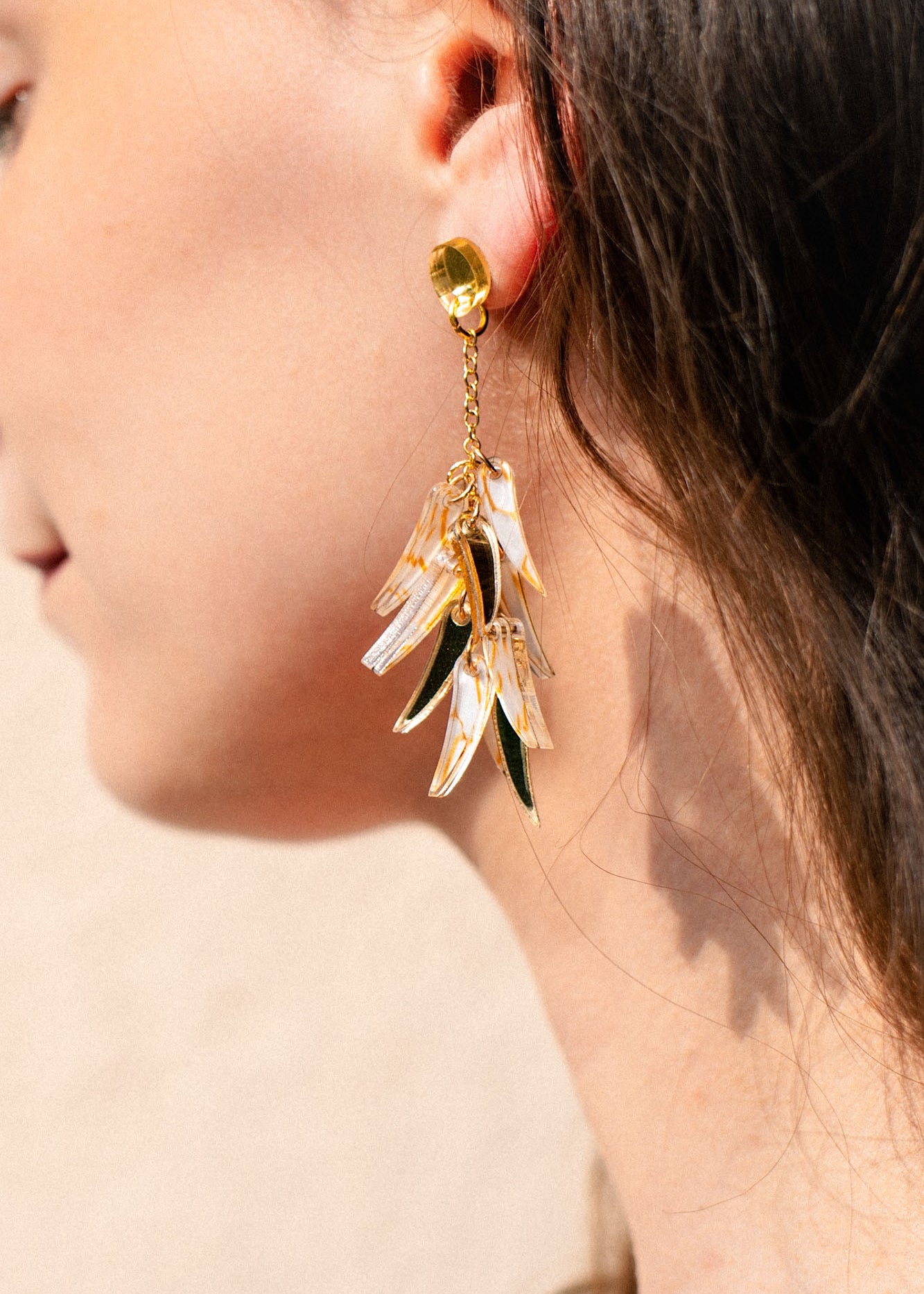 Close-up of a woman’s ear wearing featherlight dangle earrings with gold, green, and translucent acrylic tufts shaped like leaves. The softly blurred background highlights the jewelry and the side of her face and hair.