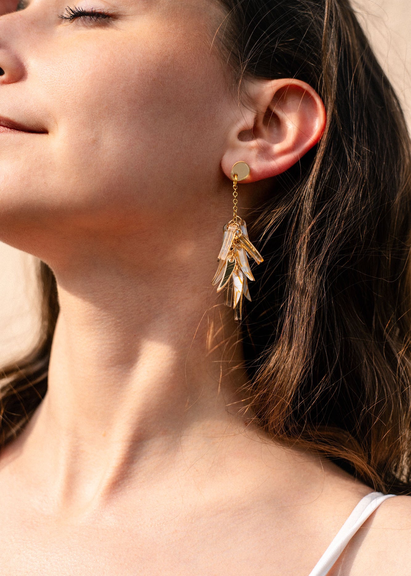 Close-up of a woman with long brown hair, eyes closed, and a slight smile, wearing featherlight gold dangle earrings shaped like cascading leaves. She is dressed in a white top as sunlight highlights her skin.