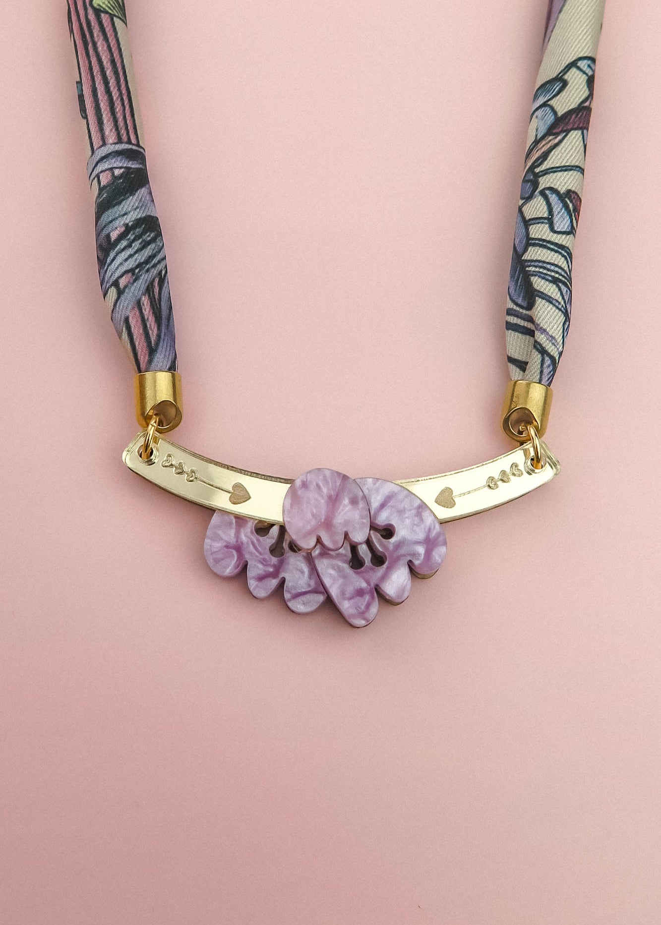 A handmade necklace featuring a pink and purple patterned fabric strap, a gold curved bar with small heart cutouts, and a cluster of marbled lavender decorative stones, displayed on a light pink background.
