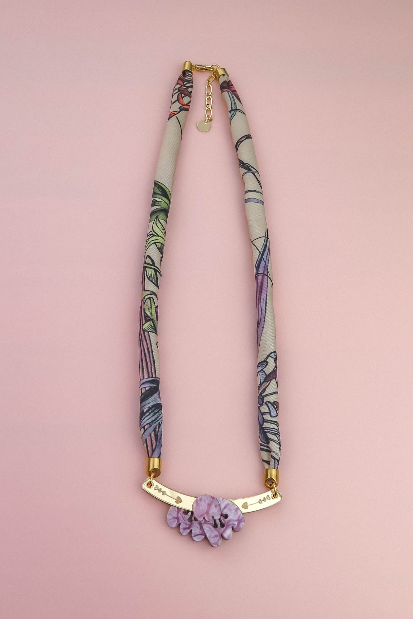 A handmade necklace with a fabric-wrapped band in muted colors and abstract patterns, featuring a gold bar centerpiece adorned with lilac stones, displayed on a light pink background—reminiscent of an elegant silk-print scarf.