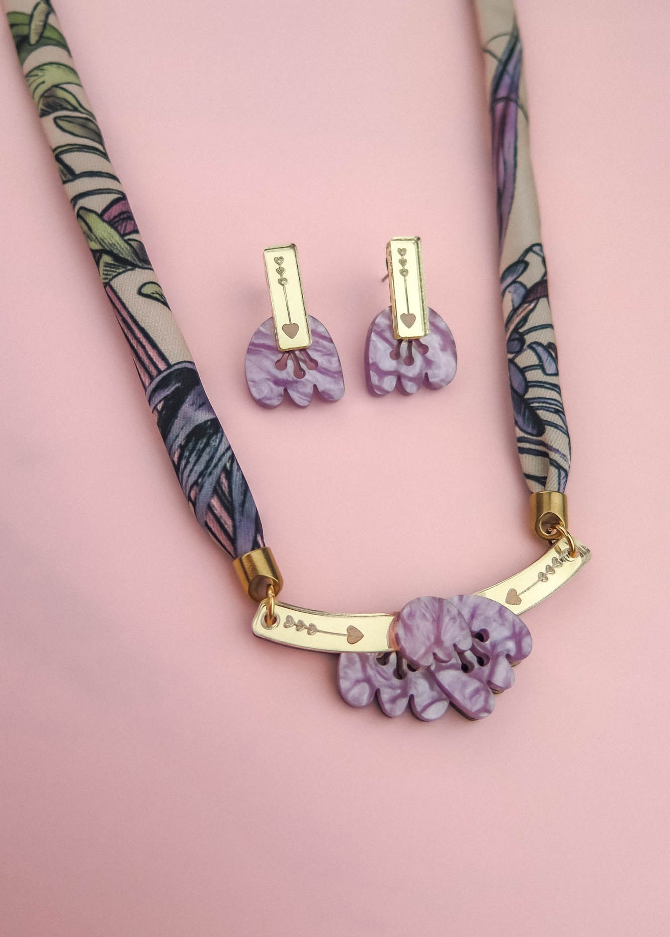 A patterned fabric handmade necklace with gold accents and a pendant featuring marbled purple shapes, paired with matching marbled purple earrings, is displayed on a soft pink background.