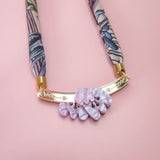 A handmade necklace with a patterned fabric band, gold metal bar, and three textured lilac stones, displayed on a pink background—perfect for pairing with an elegant silk-print scarf.