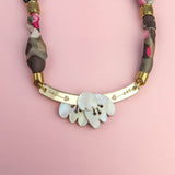 A handmade necklace with a gold curved bar, white shell-like pieces, and a multicolored fabric chain reminiscent of a silk-print scarf in green, pink, brown, and gold accents, displayed on a light pink background.