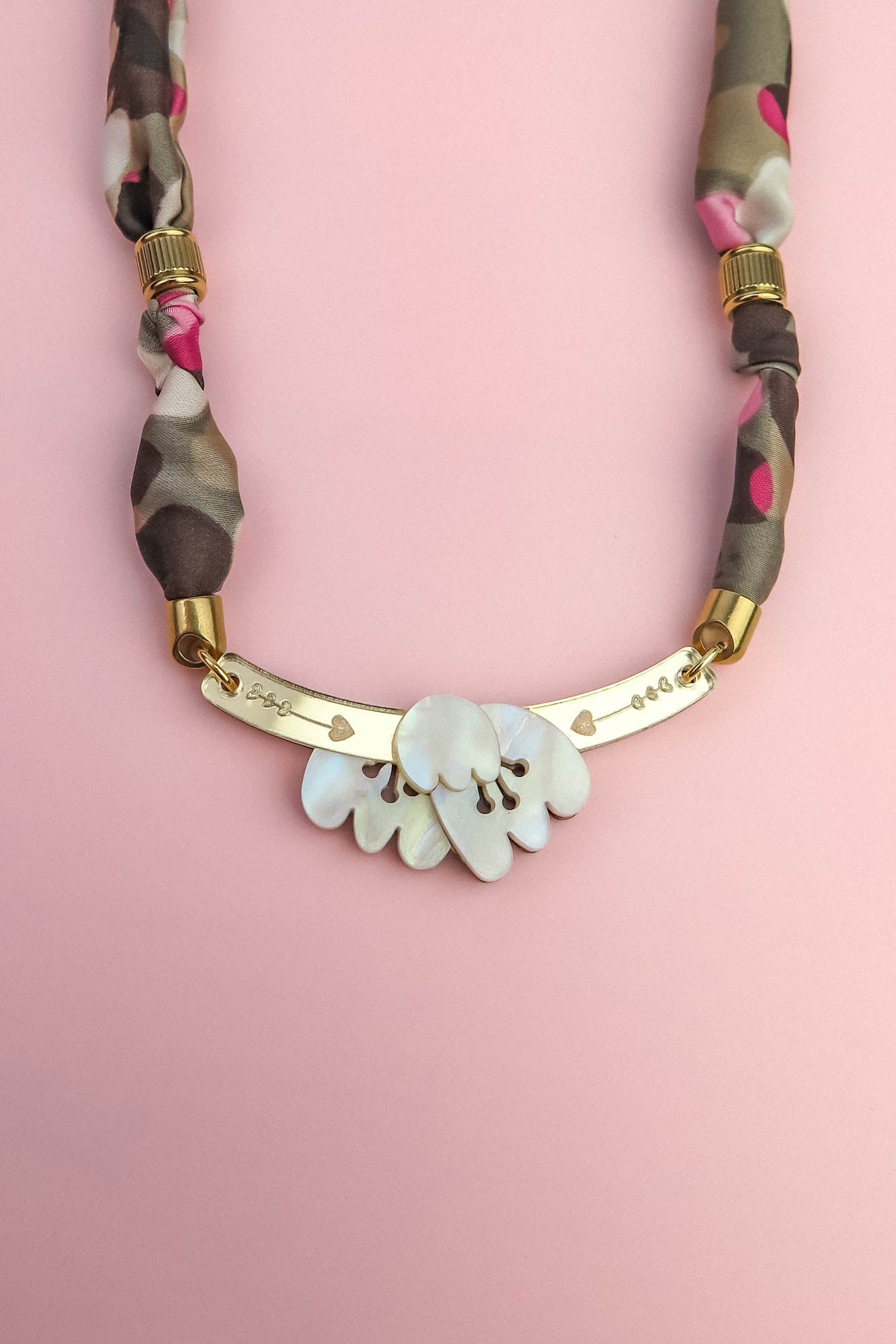 A handmade necklace with a gold curved bar, white shell-like pieces, and a multicolored fabric chain reminiscent of a silk-print scarf in green, pink, brown, and gold accents, displayed on a light pink background.