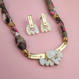 A handmade necklace and matching earrings with gold and mother-of-pearl charms are displayed on a pink background. The necklace features a patterned fabric chain, while the earrings have rectangular gold accents.