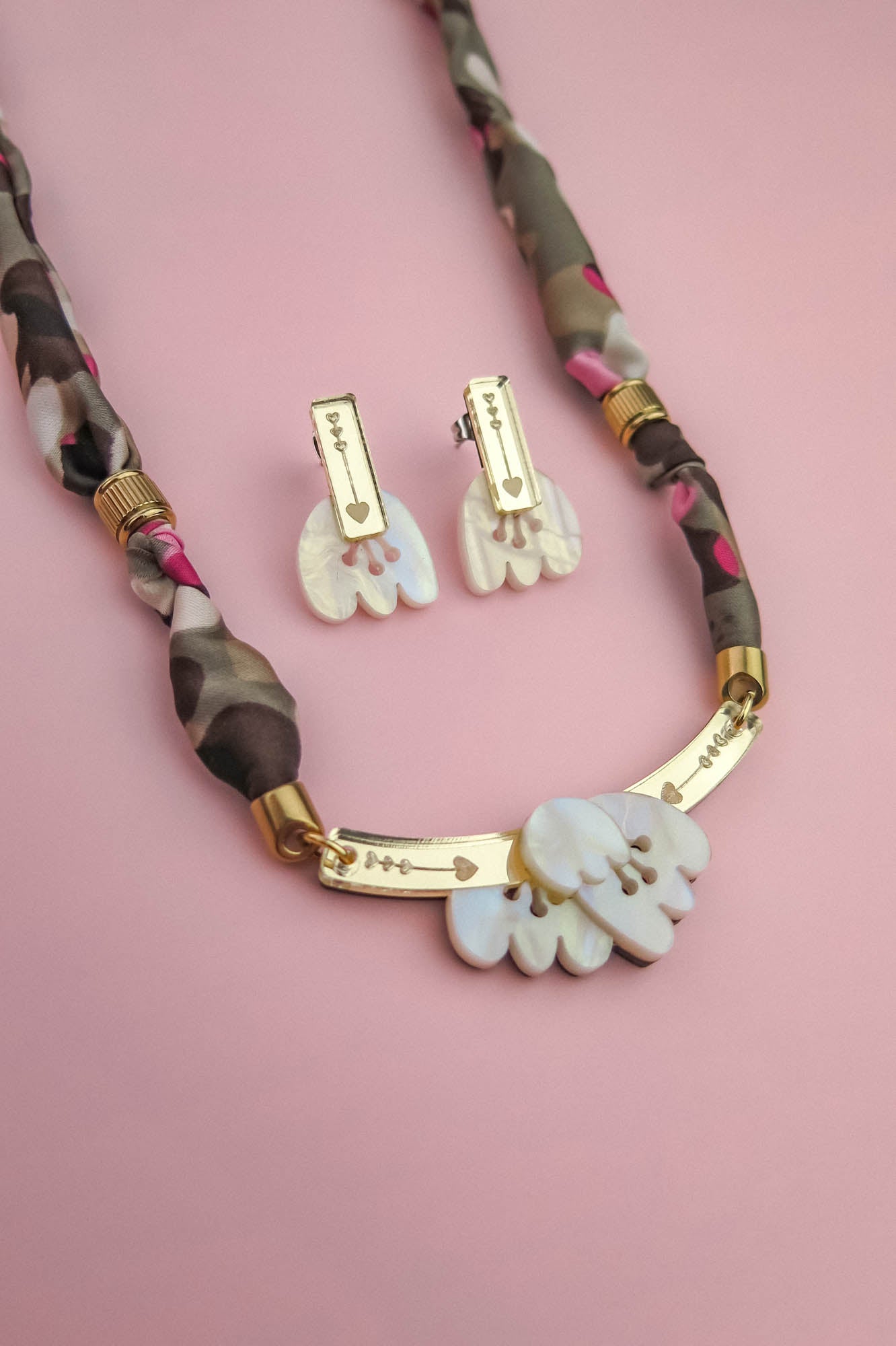 A handmade necklace and matching earrings with gold and mother-of-pearl charms are displayed on a pink background. The necklace features a patterned fabric chain, while the earrings have rectangular gold accents.