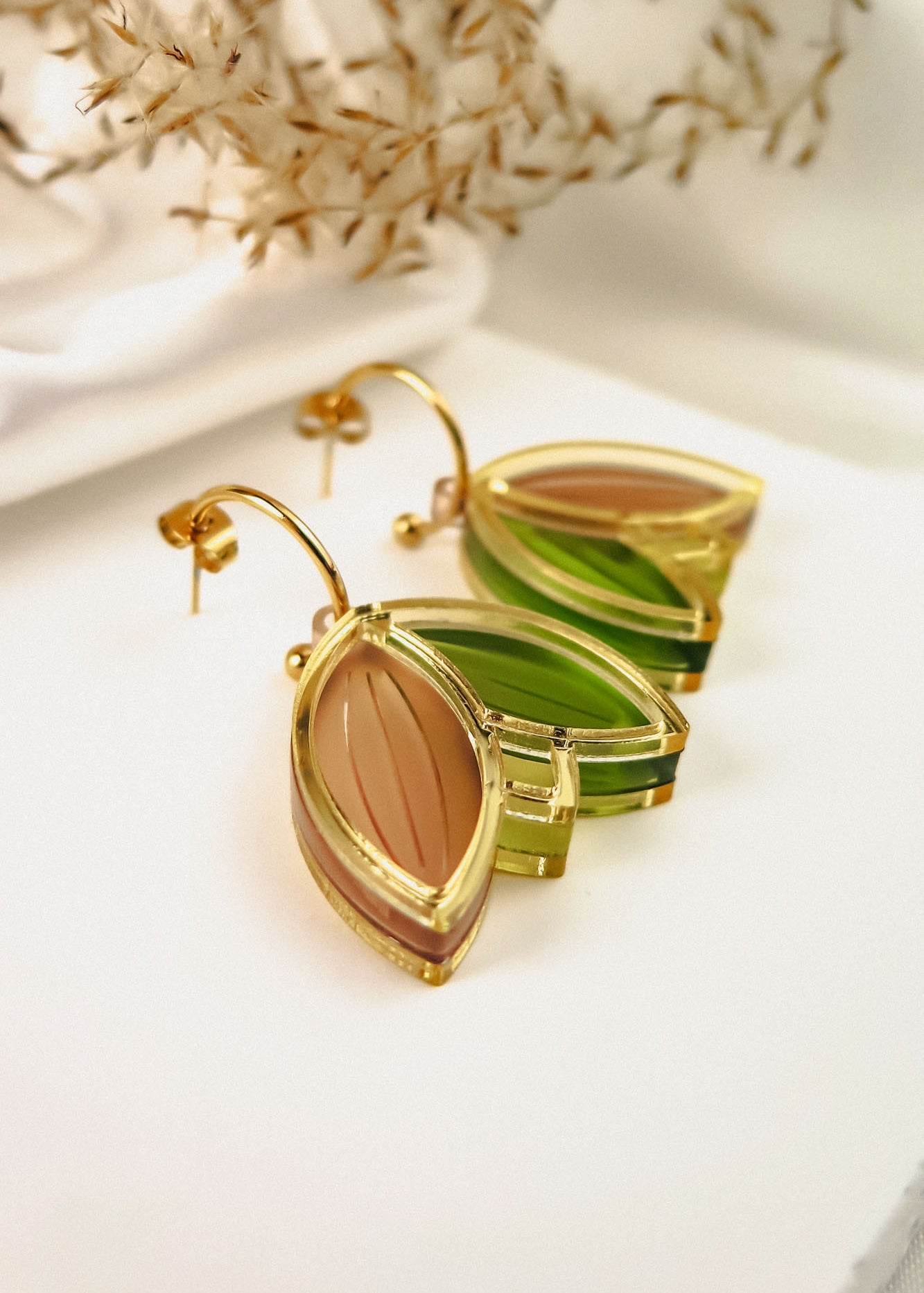 A pair of gold, leaf-shaped handcrafted earrings with translucent green and brown sections, displayed on a white surface with dried foliage in the background.