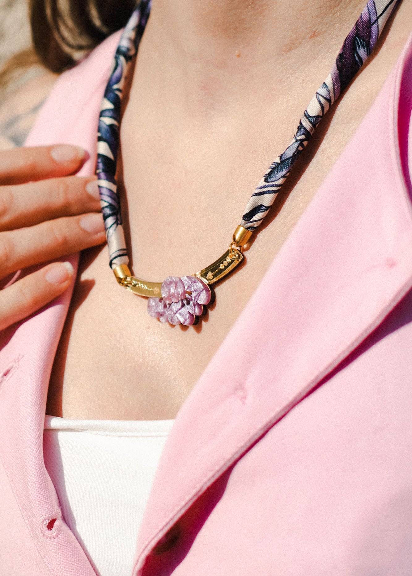 A person wearing a pink shirt and white top displays a handmade necklace with a patterned fabric band and gold clasp, featuring pink and purple gemstone flowers. Their hand rests near the collarbone, complementing an elegant silk-print scarf.