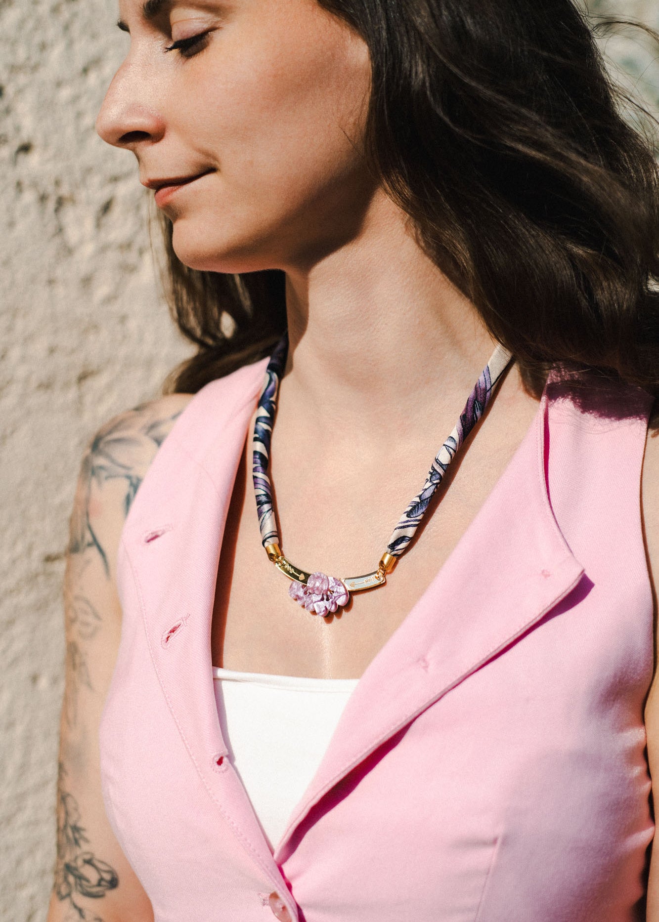 A woman with long dark hair and a floral tattoo on her arm wears a pink sleeveless vest, a white top, and a handmade necklace with a pink pendant, standing outdoors in sunlight.
