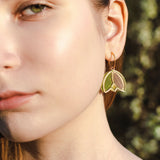 Close-up of a young woman’s face in natural light, focusing on her left eye, cheek, and ear. She wears lightweight handcrafted earrings shaped like gold and green leaves, with a blurred outdoor background.