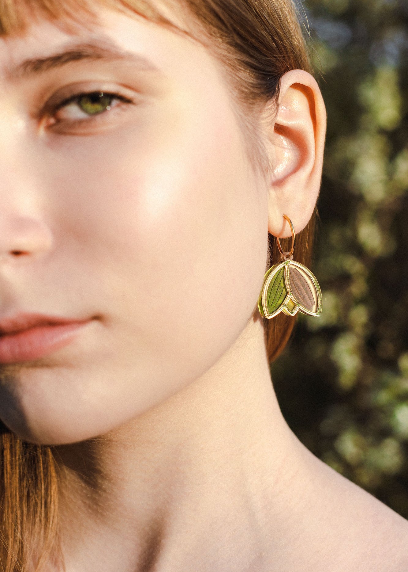 Close-up of a young woman’s face in natural light, focusing on her left eye, cheek, and ear. She wears lightweight handcrafted earrings shaped like gold and green leaves, with a blurred outdoor background.