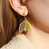 Close-up of a person’s ear wearing handcrafted Tulip Huggie Earrings with three leaf-shaped pendants in brown, green, and tan. The person has light brown hair and is dressed in a cream-colored, textured garment.